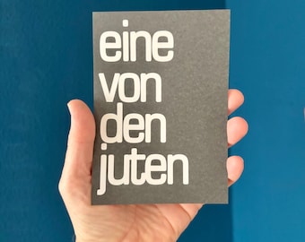 May include: A rectangular, dark gray card with the German text "eine von den juten" in white, held against a blue background. The card is held in a person's hand.