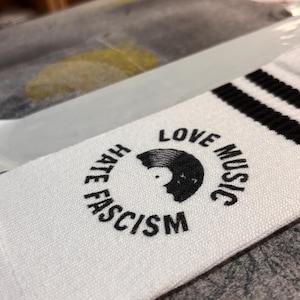 May include: A white sock featuring black stripes at the top and a circular black graphic. The design includes a vinyl record silhouette and the text "LOVE MUSIC HATE FASCISM" arranged in a circle around the central record icon.