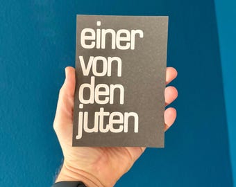 May include: A dark gray rectangular card with the German text "einer von den juten" in white, held in a hand. The card is against a blue background. A black smartwatch is visible on the wrist.