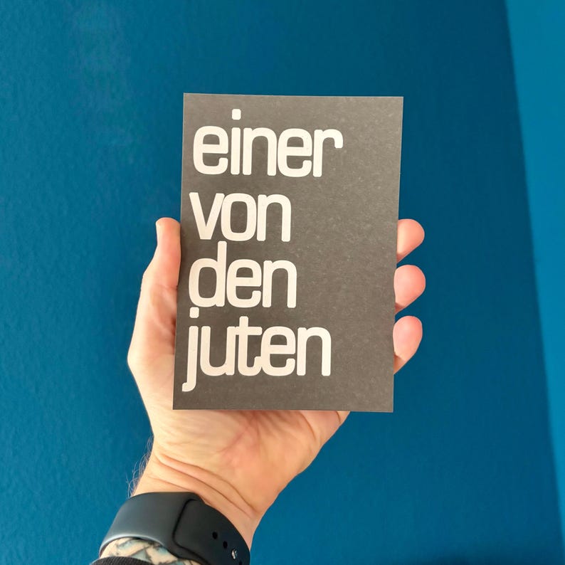 May include: A dark gray rectangular card with the German text "einer von den juten" in white, held in a hand. The card is against a blue background. A black smartwatch is visible on the wrist.