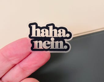 May include: A die-cut sticker featuring the text "haha. nein." in a bold, rounded, black sans-serif font. The letters have a metallic silver finish that reflects light, and the sticker has a clear border following the shape of the text.