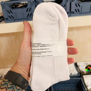 May include: A pair of plain white socks held together by a white paper band. The band features printed text including material composition of 62% organic cotton, 35% recycled polyester, and 3% elastane, with care instructions and origin details.