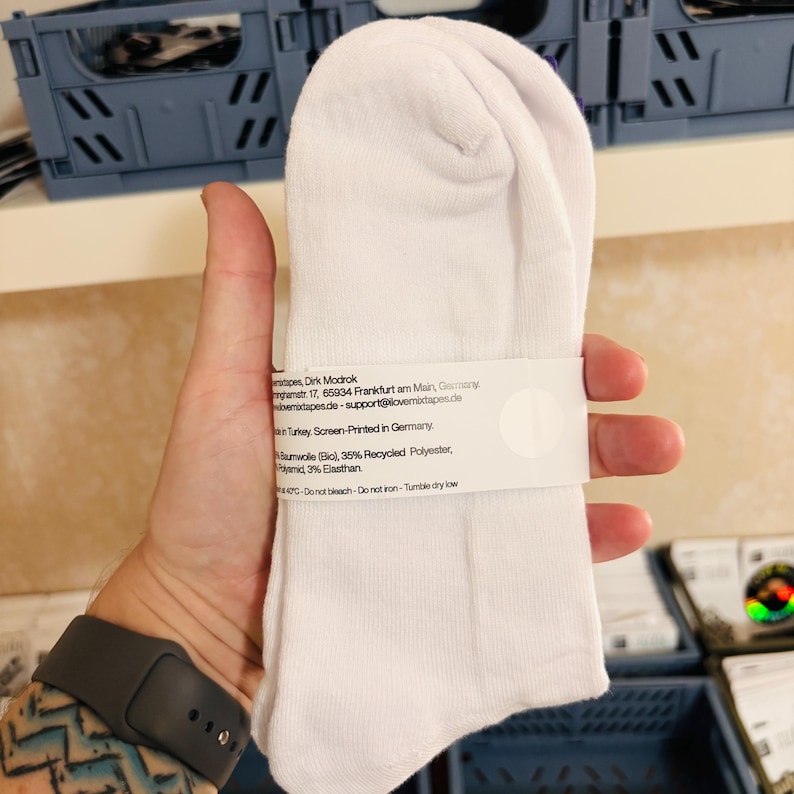 May include: A pair of plain white socks held together by a white paper band. The band features printed text including material composition of 62% organic cotton, 35% recycled polyester, and 3% elastane, with care instructions and origin details.