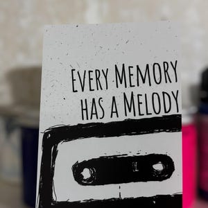 May include: A white card featuring the text "EVERY MEMORY HAS A MELODY" in black, above a black illustration of a cassette tape. The card has a speckled texture and is set against a neutral background.