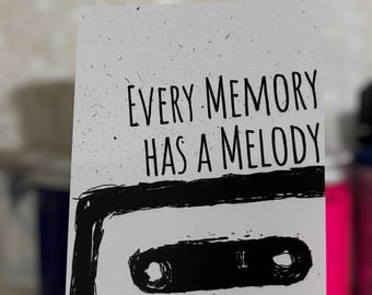 May include: A white card with the text "EVERY MEMORY HAS A MELODY" in black, above a black illustration of a cassette tape. The card has a speckled texture and is set against a neutral background.
