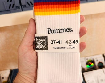 May include: White crew socks featuring a ribbed texture and a horizontal rainbow stripe pattern at the top. The word Pommes. is printed in black below the stripes. A paper band displays the text I love mix tapes, 37-41, 42-46, and SCREEN-PRINTED SOCKS.
