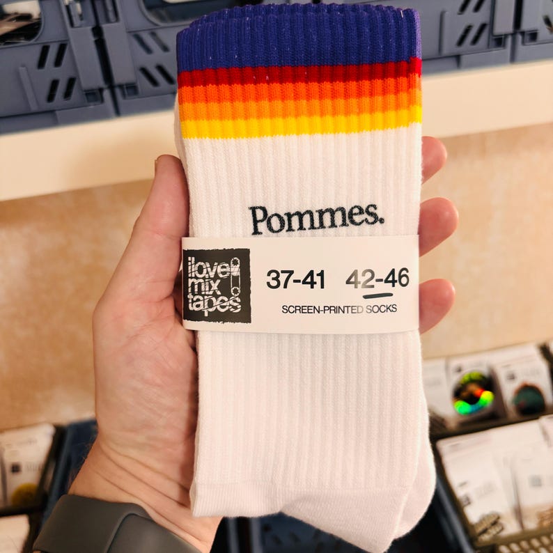 May include: White crew socks featuring a ribbed texture and a horizontal rainbow stripe pattern at the top. The word Pommes. is printed in black below the stripes. A paper band displays the text I love mix tapes, 37-41, 42-46, and SCREEN-PRINTED SOCKS.