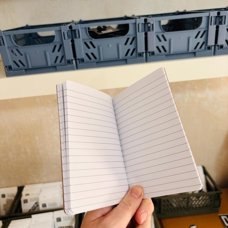 May include: An open notebook with white lined pages. The notebook features a simple binding and is held in a vertical orientation. The pages are clean and feature horizontal black lines for writing, providing a clear view of the interior paper structure.
