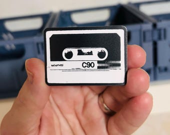 May include: A rectangular pin featuring a black and white graphic of a cassette tape. The design includes the text C90 and the word NOEMUTAPES printed on the lower section of the tape label, with a white border surrounding the black central area.