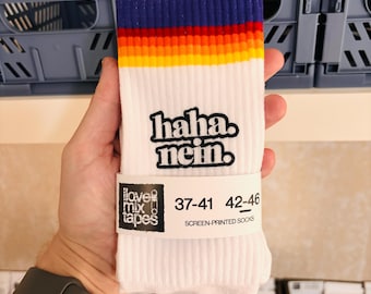 May include: White ribbed crew socks featuring a dark blue, red, and yellow striped cuff. The text "haha. nein." is embroidered in black on the front. A white paper band displays the text "ilovemixtapes" and size ranges 37-41 and 42-46.