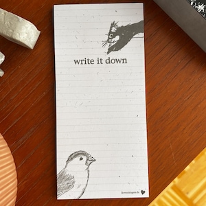 Könnte beinhalten: Ein weißer, linierter Notizblock mit dem Text "write it down" und Illustrationen eines Vogels und einer Hand mit einer Katze. Der Notizblock hat ein kleines Herzsymbol und die Website-Adresse ilovemistapes.de.