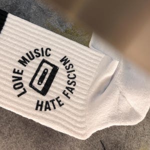 Könnte beinhalten: Eine weiße, gerippte Socke mit einem schwarzen Grafikdruck einer Kassette, umgeben von dem kreisförmigen Text LOVE MUSIC HATE FASCISM. Die Socke hat einen schwarzen Streifen am oberen Rand und eine deutlich sichtbare, gerippte Strickstruktur.