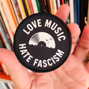 Könnte beinhalten: Ein kreisförmiger, schwarzer, bestickter Aufnäher mit weißem Rand. In der Mitte befindet sich eine weiße Grafik einer Schallplatte mit zwei kleinen Vögeln. Der Text "LOVE MUSIC" ist oben und "HATE FASCISM" unten in weißen Großbuchstaben gebogen.
