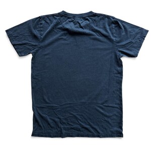 May include: A navy blue, short-sleeved t-shirt, laid flat on a white surface. The shirt has a crew neck and appears to be made of a soft, worn material. The fabric has a slightly faded appearance, suggesting it has been washed.