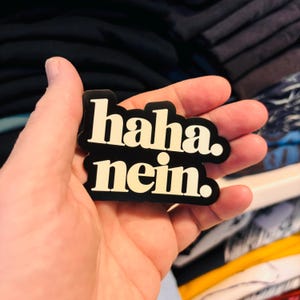 Könnte beinhalten: Ein schwarzer, konturgestanzter Aufkleber mit cremefarbener serifenloser Schrift, auf dem "haha. nein." steht. Der Aufkleber hat eine glatte, matte Oberfläche und ein kräftiges, blockartiges Design, bei dem der Text in zwei Zeilen angeordnet ist.
