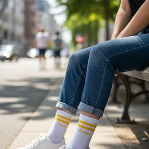 May include: White crew socks featuring two horizontal yellow stripes near the top and the word Pommes printed in yellow text. The socks are paired with blue denim jeans that have a cuffed hem and clean white low-top sneakers with white laces.