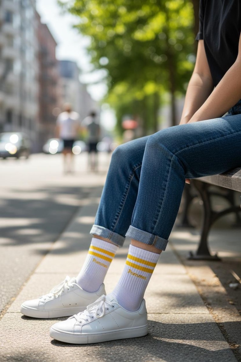 May include: White crew socks featuring two horizontal yellow stripes near the top and the word Pommes printed in yellow text. The socks are paired with blue denim jeans that have a cuffed hem and clean white low-top sneakers with white laces.