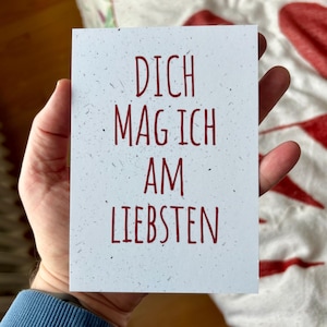 May include: A rectangular white greeting card with a speckled texture featuring the red text DICH MAG ICH AM LIEBSTEN printed in a simple sans-serif font centered on the front of the card.