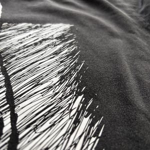 May include: Close-up of a dark gray t-shirt with a white graphic design. The design features a series of black, scribbled lines against a white background. The fabric appears soft and slightly wrinkled.