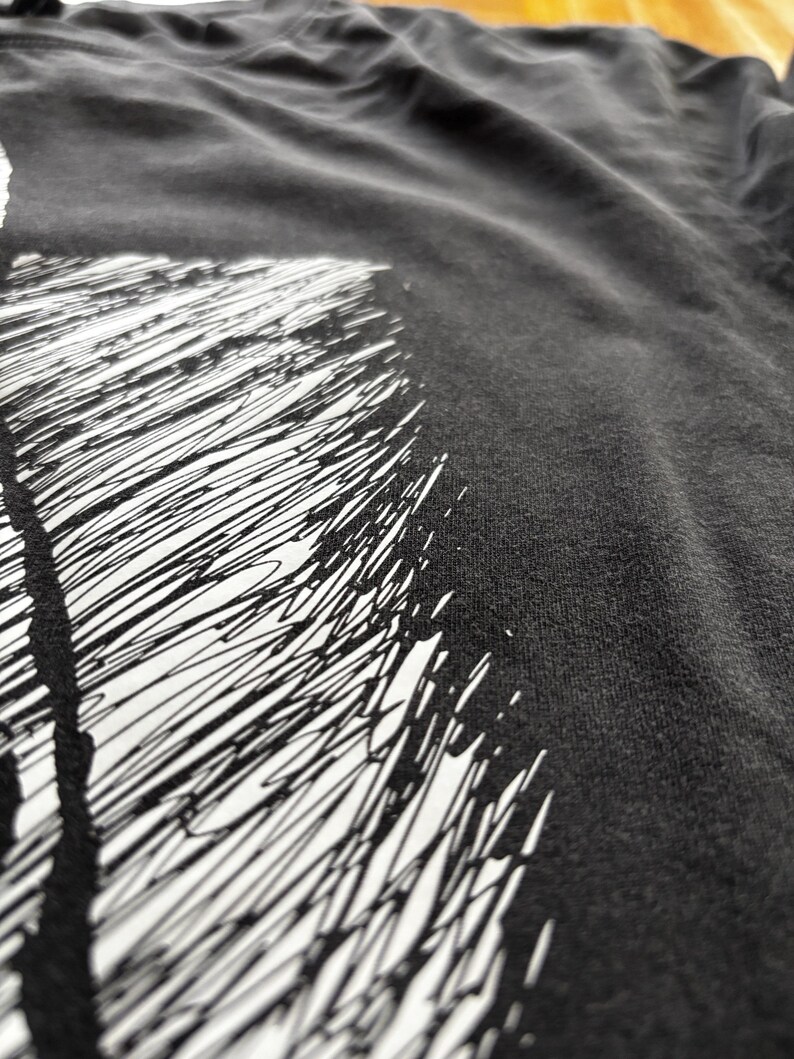 May include: Close-up of a dark gray t-shirt with a white graphic design. The design features a series of black, scribbled lines against a white background. The fabric appears soft and slightly wrinkled.