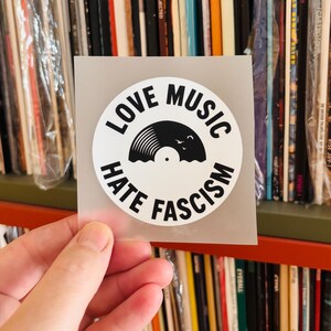 Könnte beinhalten: Ein runder weißer Aufkleber mit schwarzem Text, der LOVE MUSIC HATE FASCISM lautet. In der Mitte befindet sich eine schwarze Grafik einer Schallplatte mit einer kleinen Silhouette von Vögeln darüber, auf einer halbtransparenten quadratischen Trägerfolie.