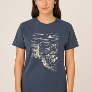 Pu&ograve; includere: T-shirt blu navy con un disegno di paesaggio color crema. Il disegno raffigura una costa con rocce, acqua e un sole o una luna. Il design crea un contrasto con il blu scuro della maglietta.