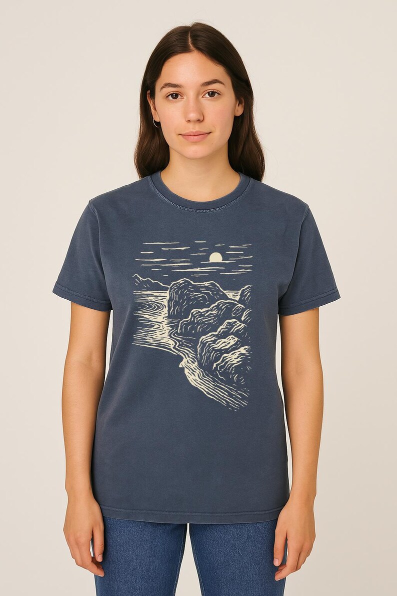 Pu&ograve; includere: T-shirt blu navy con un disegno di paesaggio color crema. Il disegno raffigura una costa con rocce, acqua e un sole o una luna. Il design crea un contrasto con il blu scuro della maglietta.