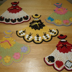May include: Five crocheted doilies featuring a woman in a dress with different designs. The doilies are white with colorful accents. One doily features a woman with a red dress and black Mickey Mouse heads around the edge. Another doily features a woman with a yellow dress and bees around the edge. Another doily features a woman with a pink dress and butterflies around the edge. Another doily features a woman with a green dress and flowers around the edge. The last doily features a woman with a red dress and playing cards around the edge.