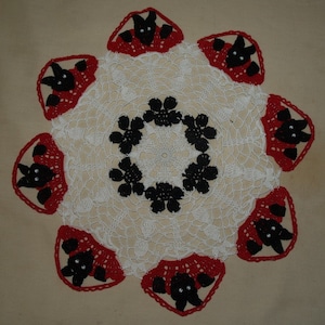 May include: A handmade, white crocheted doily with a circular design. The doily features a central ring of black floral shapes and is bordered by red and black bat-shaped accents. The bats have small, sparkly eyes.