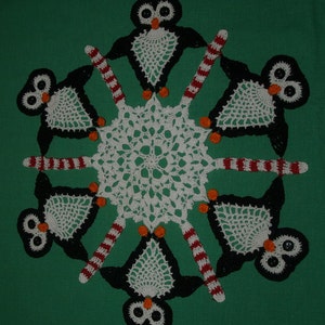 May include: A white crocheted snowflake doily with six black and white crocheted penguins around the edge. The penguins are holding red and white striped candy canes.