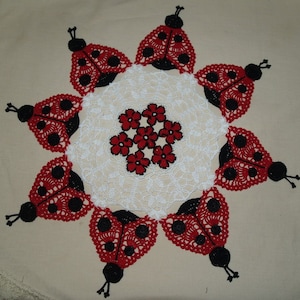 May include: A white crocheted doily with a red and black ladybug design. The doily is circular and has a scalloped edge. The ladybugs are arranged in a circle around the center of the doily.