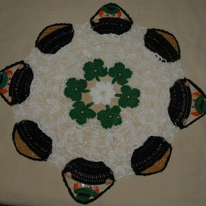 May include: White crocheted doily with a green shamrock design in the center. The doily is decorated with black and gold crocheted leprechaun figures around the edge.
