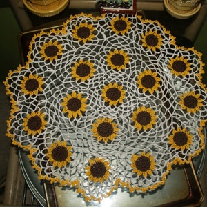 May include: A white crocheted doily with a sunflower design. The doily is made with a delicate crochet pattern and features brown and yellow sunflowers.