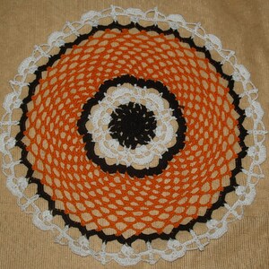 May include: A crocheted doily with a black, white, and orange design. The doily has a circular shape and is made with a delicate, lacy pattern.