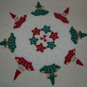 Puede incluir: Doily de crochet blanco con un patrón de estrellas rojas y verdes en el centro. Ocho figuras de crochet de elfos y Papá Noel rodean el doily.