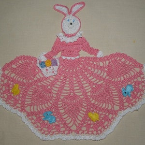 May include: Pink crocheted doily with a white crocheted bunny in the center holding a basket of colorful eggs. The doily is decorated with crocheted bunnies and flowers.