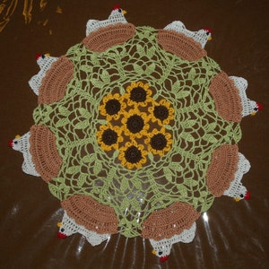 May include: A crocheted doily with a green centre and brown accents. The doily is decorated with a sunflower design and eight crocheted chickens around the edge.