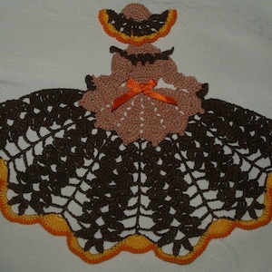 May include: A brown crocheted doily with an orange trim. The center of the doily features a crocheted doll with a brown dress and an orange bow.