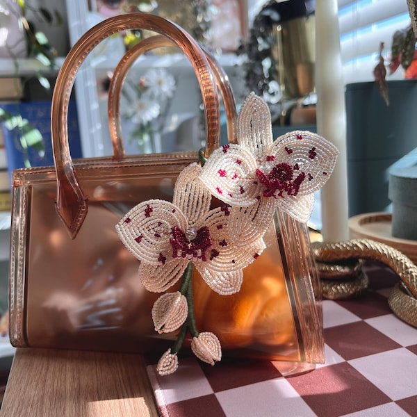 Pode incluir: Uma mala de mão translúcida em tom de cobre com duas pegas e um acessório decorativo de orquídea feita de missangas. A orquídea é composta por missangas cor de creme com detalhes em vermelho escuro nas pétalas e caules verdes com botões.