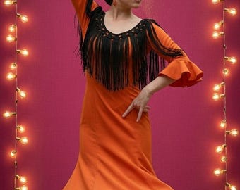 Op de afbeelding: Een vrouw in een oranje flamencojurk met zwarte franjes, versierd met bloemen in haar haar, danst. De jurk heeft uitlopende mouwen en een zwierige rok. Slingers omlijsten de danseres tegen een magenta achtergrond.