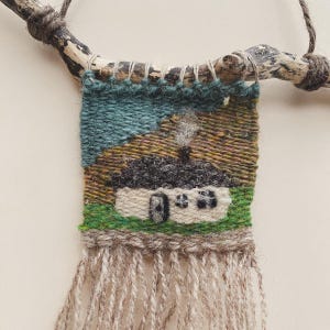 May include: A woven tapestry depicting a small cottage with a smoking chimney, set against a backdrop of green and blue hues. The artwork is suspended from a natural wooden branch, with fringed yarn at the bottom.