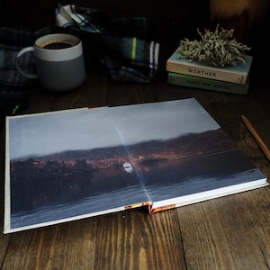 May include: An open hardcover book displays a full-page photograph of a boat on a dark, misty lake surrounded by mountains. Nearby are a two-toned gray and white ceramic mug, a plaid fabric, two stacked books, and a wooden pencil.