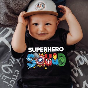 A smiling baby wearing a black t-shirt with superhero designs and a white baseball cap.