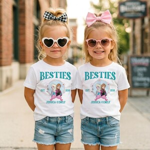 Two young girls wearing matching white t-shirts with the text "BESTIES" and "BESTIES" printed on them, along with a graphic of two girls, are standing side by side on a city street.