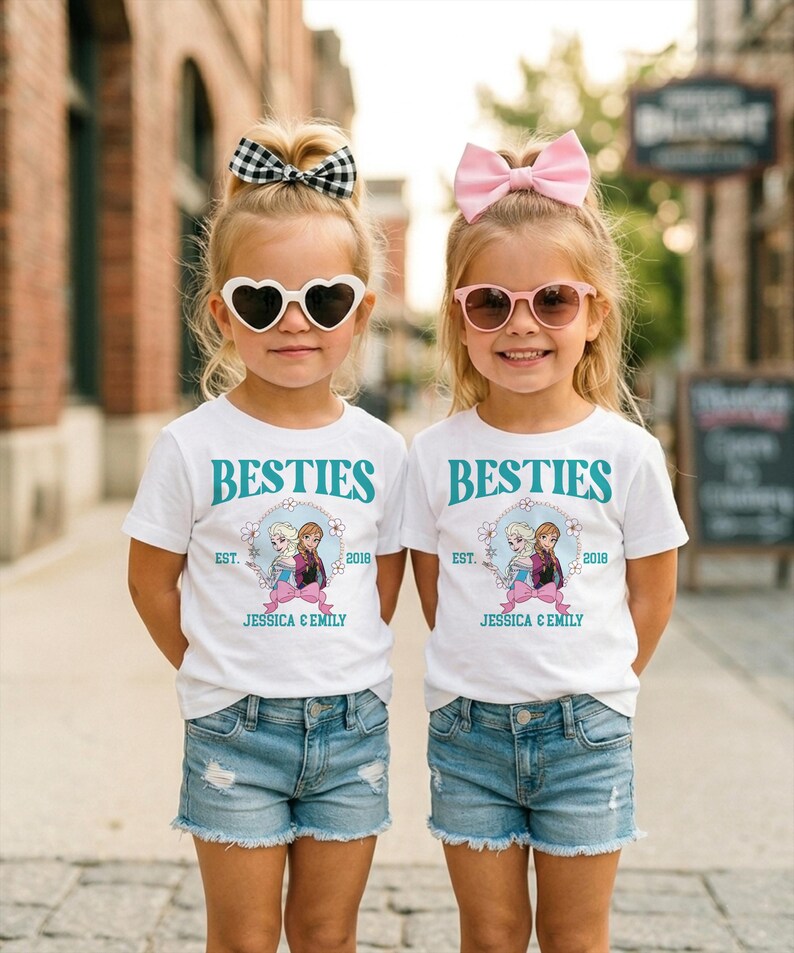 Two young girls wearing matching white t-shirts with the text "BESTIES" and "BESTIES" printed on them, along with a graphic of two girls, are standing side by side on a city street.