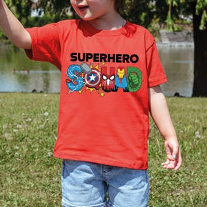 A young child wearing a red t-shirt with the word "SUPERHERO" printed on it, standing in a grassy field near a body of water.
