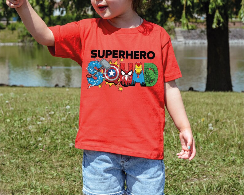 A young child wearing a red t-shirt with the word "SUPERHERO" printed on it, standing in a grassy field near a body of water.