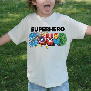 A young child wearing a white t-shirt with the text "SUPERHERO" printed on it, surrounded by various superhero logos and characters.