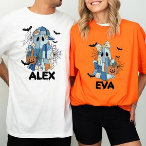 May include: White and orange Halloween t-shirts with matching ghost designs. Each ghost is patchwork-style, holding a pumpkin and surrounded by bats and spiderwebs. The names "ALEX" and "EVA" are printed below the ghosts.