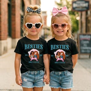 Two young girls wearing matching black t-shirts with the text "BESTIES" and "BESTIES" printed on them, along with a heart-shaped logo, are standing together on a city street.
